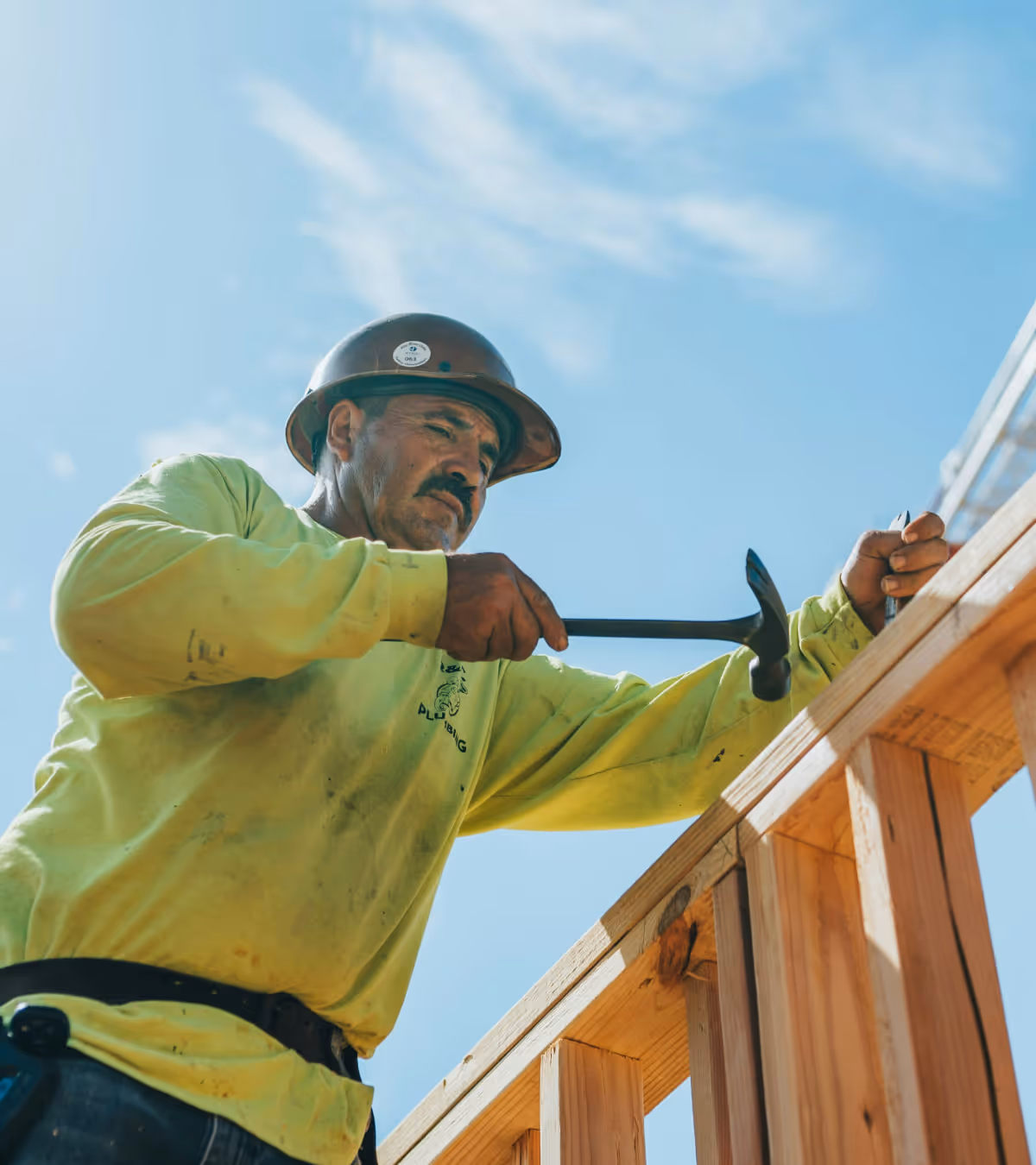 Construction worker in a yellow shirt and hard hat hammering a nail into a wooden frame under a blue sky.