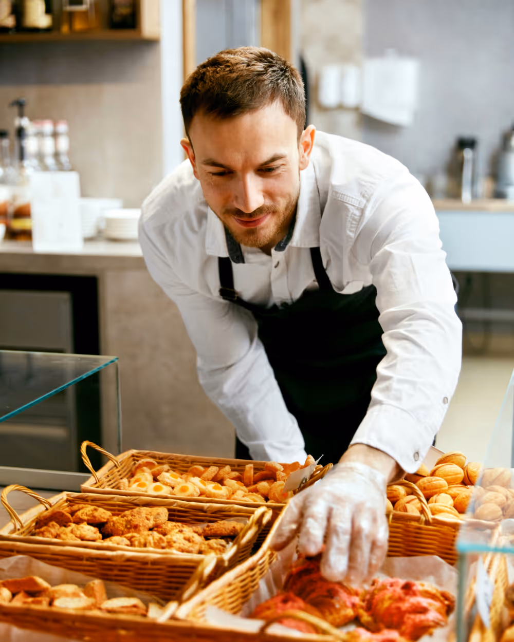 Male bakery worker in a white shirt and black apron arranging assorted pastries in wicker baskets.